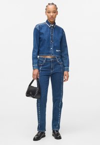 Set di giacca in denim e jeans in blu scuro, caratterizzato da borchie argentate sul colletto e sulle cuciture laterali. Include una borsa nera con dettagli in borchie.