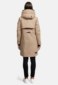 Parka isolée beige avec une grande capuche, un rabat arrière surdimensionné et des détails en noir. Elle comporte une fermeture éclair et un écusson de marque près de l'ourlet.