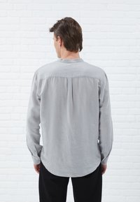 Lichtgrijs langemouwen shirt met een mandarinokraag, plooidetails aan de achterkant en knoopsluiten op de manchetten, gemaakt van een zachte, textuurrijke stof.