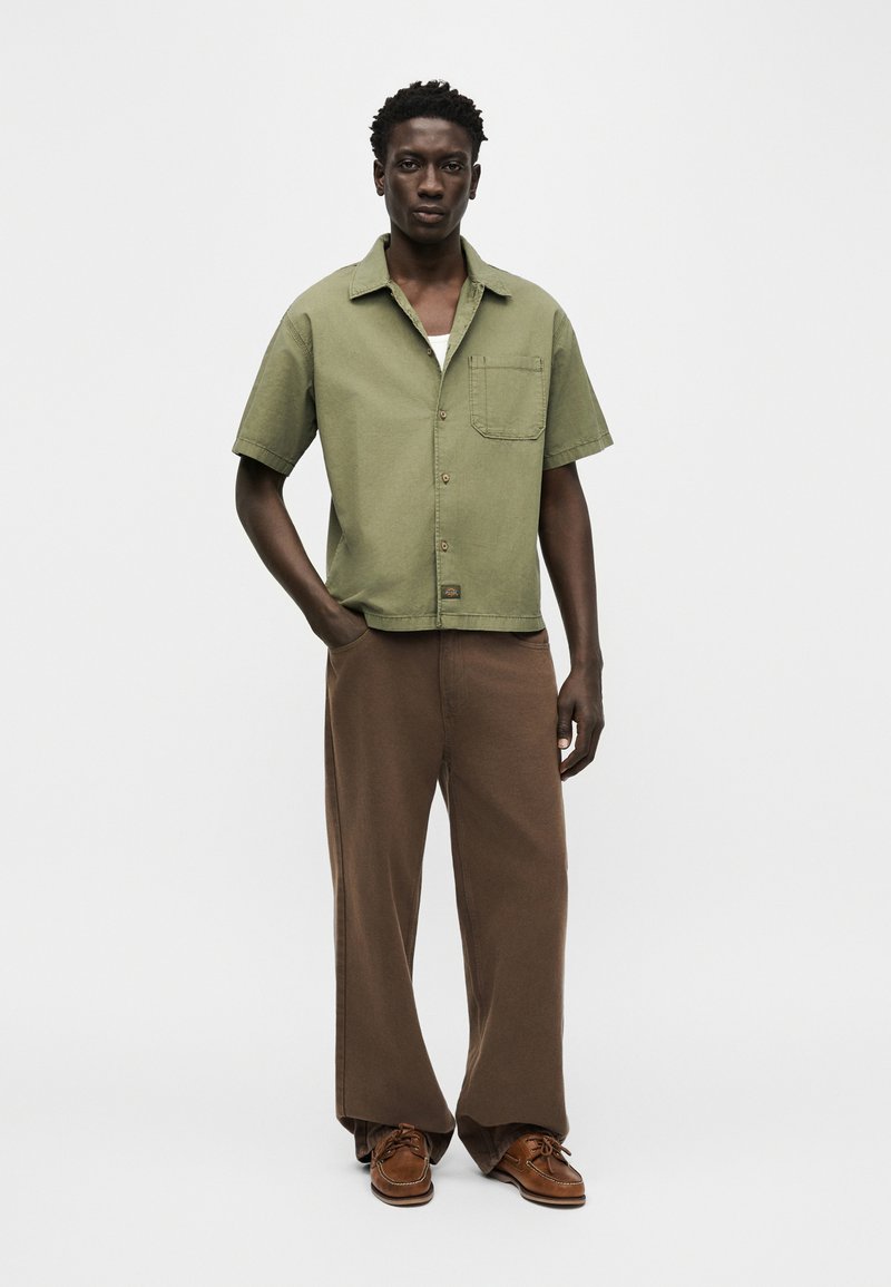 Hombre de pie con las manos en los bolsillos, vestido con una camisa verde de manga corta con botones, pantalones anchos marrones y zapatos marrones.