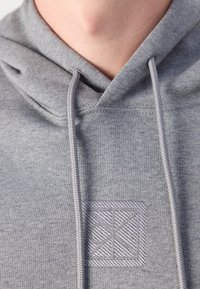 Gros plan d'un sweat à capuche gris avec cordons et un logo carré brodé en relief sur la poitrine, porté par une personne sans visage visible.
