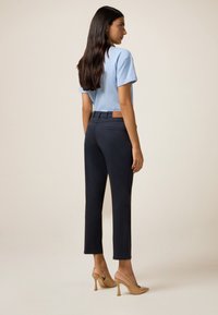Pantalons navy à coupe slim avec une poche arrière, associés à une chemise à manches courtes bleu clair et des chaussures à talons hauts nude. Fond uni.