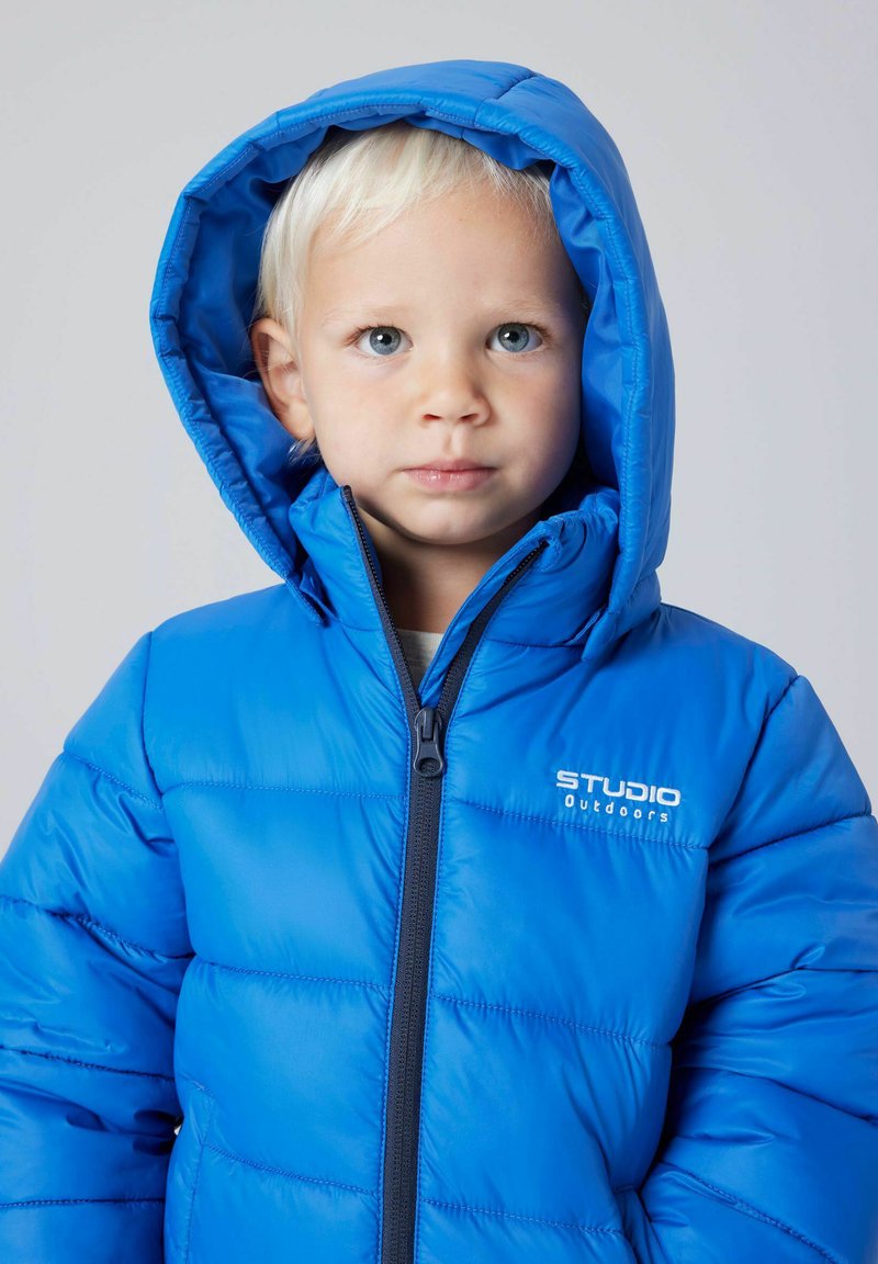 Jeune enfant aux cheveux blonds clairs portant une doudoune à capuche bleue avec le logo "Studio Outdoors" sur la poitrine sur un fond uni.
