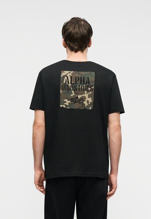 Muž v černém tričku s maskáčovým čtvercem a nápisem "ALPHA INDUSTRIES" na zádech, stojící před jednoduchým světlým pozadím.