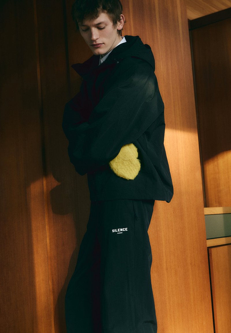 Black outerwear with a large yellow textured patch on the sleeve, paired with black pants featuring "SILENCE" text in white.