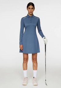 Sininen golf-mekko poolokauluksella, pitkillä hihoilla ja levenevällä helmalla. Olkapäillä valkoiset raidat. Yhdistetty golf-hansikkaaseen ja mailaan.