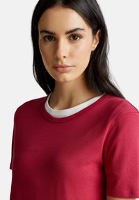 Giovane donna con capelli lunghi e scuri che indossa una maglietta rossa a maniche corte sovrapposta a una camicia bianca a girocollo, rivolta in avanti con un'espressione neutra.