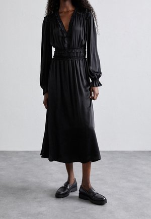 Vrouw draagt een lange zwarte satijnen jurk met gebloemde lange mouwen en zwarte loafers, staand op een grijze vloer tegen een witte muur.