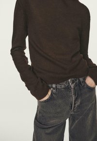 Persona che indossa una maglia a maniche lunghe marrone scuro con le mani nelle tasche di jeans di denim nero scolorito a vita alta, su uno sfondo semplice.