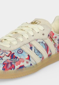 Λευκό αθλητικό παπούτσι Adidas Samba με κόκκινο και μπλε floral σχέδιο, μπεζ σουέντ λεπτομέρειες και καουτσούκ σόλα.