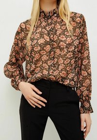 Doorzichtige blouse met lange mouwen en hoge kraag, met een donker bloemenpatroon in tinten roze en oranje, gecombineerd met een zwarte broek.
