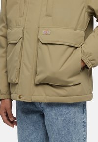 Beige isolerad jacka med två stora framsnittfickor, elastiska ärmslut och tryckknapp. Bärs över ljusblå denim. Avslappnad design.