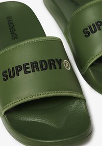 Groene schuifsandalen van glad synthetisch materiaal met een getextureerde voetbed. Voorzien van een brede band met "SUPERDRY" in het zwart gedrukt.