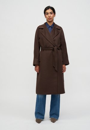 Femme portant un long manteau marron ceinturé sur une chemise bleue et un jean à jambes larges, debout devant un fond clair uni.