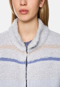 Fuzzy, light grey jacket with a high collar, featuring horizontal stripes in beige and shades of blue. Soft texture with a zip closure.