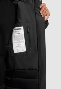 Zwarte pufferjack met gewatteerd textuur, interne ritszak en waslabel. De stof is mat met een gladde afwerking.