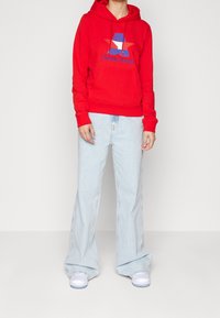 Felpa rossa con cappuccio caratterizzata da un logo con una stella e il testo "Tommy Jeans". Abbinata a jeans chiari a zampa d'elefante e sneakers bianche.