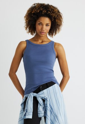 Jonge vrouw met krullend haar, gekleed in een blauwe mouwloze top en een gestreept overhemd om haar middel gestrikt, staand tegen een effen achtergrond.