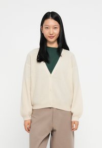 Cardigã creme com um decote em V profundo, textura canelada e botões frontais, combinado com uma blusa verde escura e calças bege.