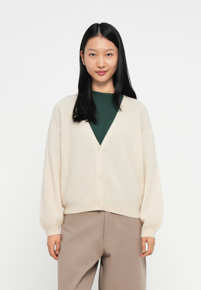 Cardigã creme com um decote em V profundo, textura canelada e botões frontais, combinado com uma blusa verde escura e calças bege.