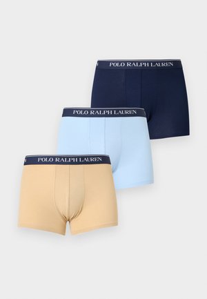 Izmos, sötét bőrű férfi áll mezítláb egyszínű háttér előtt, bézs színű Polo Ralph Lauren boxeralsót viselve.