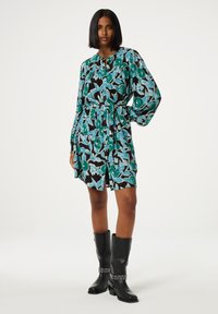 Blumenkleid mit schwarzem Grund und türkisfarbenen sowie grünen Mustern, langen Ärmeln, einem Bindegürtel in der Taille und einer weichen Textur, kombiniert mit kniehohen schwarzen Stiefeln.