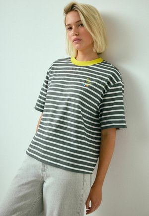 Tânără cu păr blond, purtând un tricou cu dungi gri și albe, cu guler galben, și blugi gri deschis, sprijinită de un perete alb.