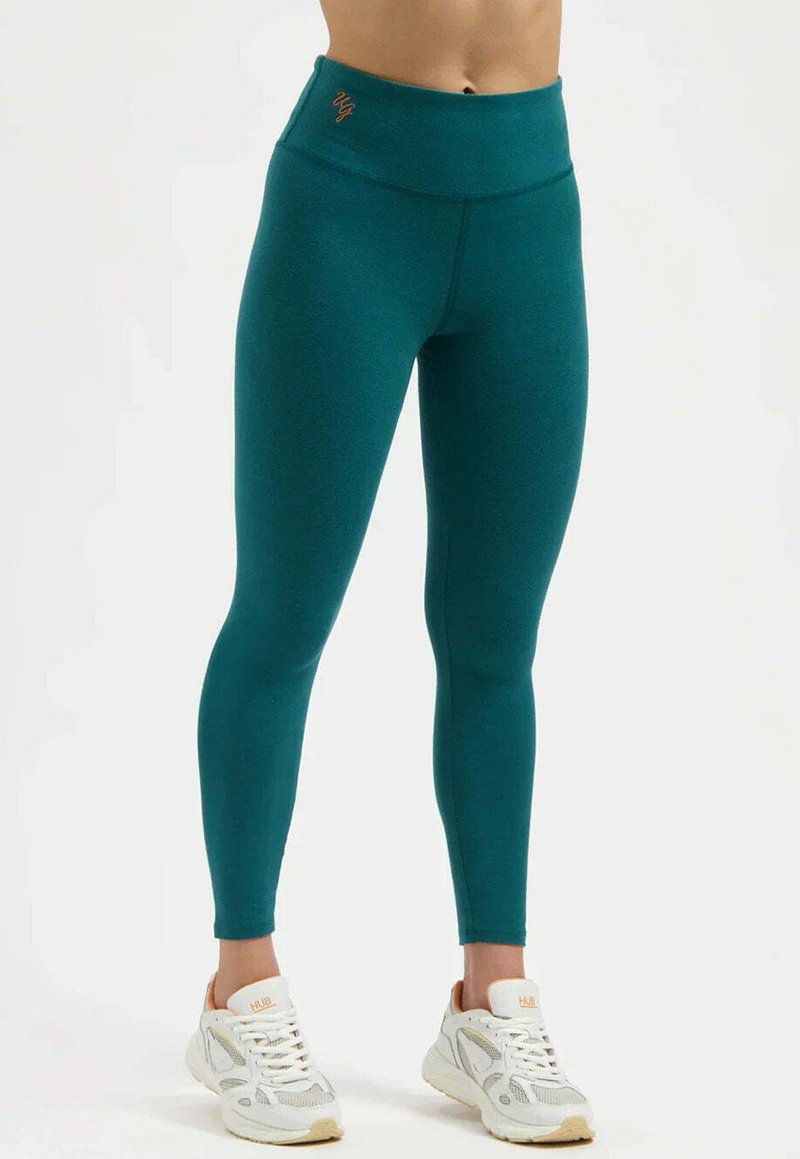 Leggings taille haute couleur sarcelle en tissu extensible, avec une texture lisse et un discret logo sur la taille, associés à des baskets blanches.