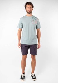 T-shirt en coton bleu clair avec un motif floral sur l'épaule gauche, associée à un short cargo violet foncé et des baskets noires avec une semelle blanche.