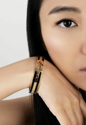 Gold and black bangle bracelet featuring a keyhole design and the "FURLA" logo, worn on a wrist with smooth skin texture.
