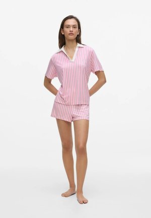 Nő áll mezítláb, rózsaszín és fehér, függőleges csíkos rövid ujjú ingben és hozzá illő rövidnadrágban, fehér háttér előtt.