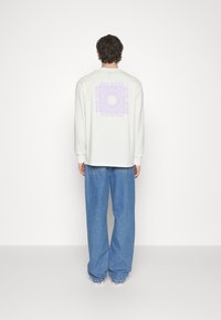 Man in een witte lange mouwen shirt met paarse 'Dickies Hard Work' graphic op de rug, losse blauwe jeans en witte sneakers, staande met zijn rug naar de kijker gekeerd.