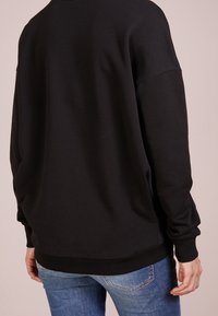 Sudadera negra de corte holgado, con puños y dobladillo acanalados, y una textura suave. Llevada con jeans azules, mostrando la vista trasera.