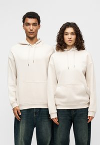 Μπεζ φούτερ με κουκούλα από απαλό ύφασμα, με τσέπη καγκουρό και κορδόνι. Συνδυασμένα με άνετο, μπλε τζιν.