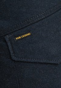 Tamno plava traper tkanina s aplikacijskom džepu koji ima crnu oznaku s natpisom "PME LEGEND" u žutom tekstu. Glatka tekstura s suptilnim šavovima.