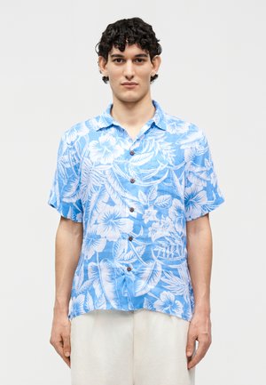 Νεαρός άνδρας με μπλε και λευκό κοντομάνικο πουκάμισο με λουλουδάτο μοτίβο και ανοιχτόχρωμο παντελόνι, στέκεται μπροστά σε απλό φόντο.