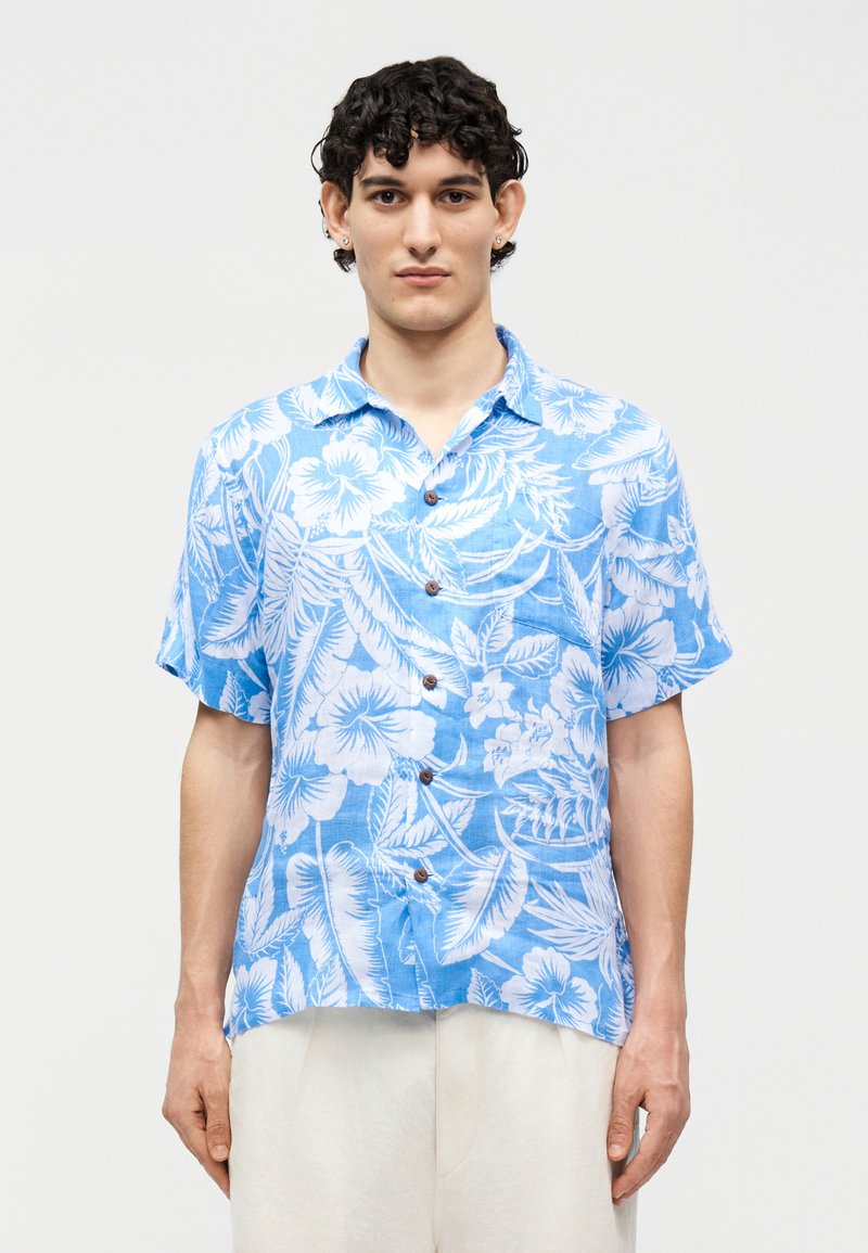 Jonge man die een blauw-wit bloemenhemd met korte mouwen en lichtgekleurde broek draagt, staand tegen een effen achtergrond.