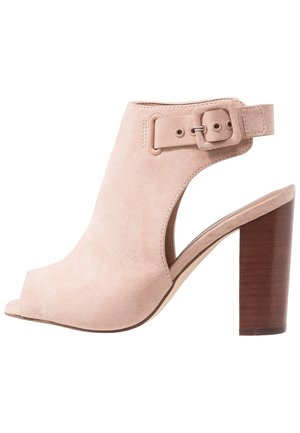 Sandale à talon ouverte en suédine beige avec un design découpé, une large bride de cheville avec boucle et un talon en bois épais.