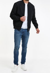 Sort bomberjakke med lynlås foran, ribbede cuffs og en glat overflade, båret over en hvid t-shirt og parret med blå jeans og hvide sneakers.
