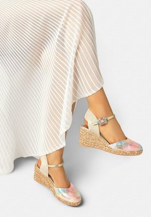 Piernas con una falda blanca plisada y sandalias de cuña de esparto en colores pastel con tiras en el tobillo y hebillas decorativas.