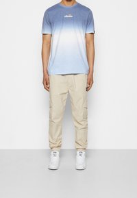 Camiseta azul claro con ombre y logo en el pecho, combinada con pantalones cargo beiges y zapatillas blancas. Atuendo simple y casual con un diseño funcional.