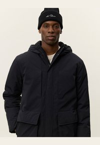 Parka de marina con capucha, con cierre de cremallera en la parte delantera, dos grandes bolsillos frontales y una textura suave. Se lleva con un gorro de punto negro.
