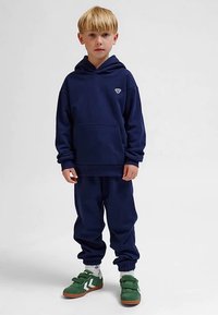 Mörkblå huvtröja med en känguruficka och liten logotyp, kombinerad med matchande joggingbyxor och gröna sneakers med kardborreband.