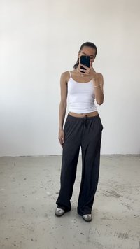 Weißes, enges Tanktop kombiniert mit lockeren, dunkelgrauen gestreiften Hosen und grauen Schlüpfschuhen. Glatte Textur und entspanntes Design.