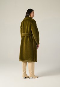 Cappotto verde oliva, morbido al tatto, con vita a cintura e maniche lunghe, abbinato a stivali beige al ginocchio e accessori minimalisti.