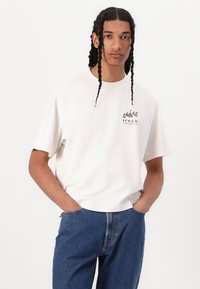 Cotton crew neck t-shirt in off-white with a small multicolored graphic reading "HVAR CROATIA" on the chest, paired with blue denim jeans.