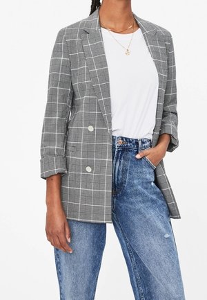 Woman wearing gray plaid double-breasted blazer over white t-shirt, paired with high-waisted blue jeans, hand in pocket.