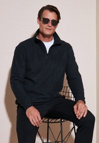 Pullover zip nera in morbido materiale, con colletto alto e dettagli della cucitura sul petto, abbinato a pantaloni neri e occhiali da sole.