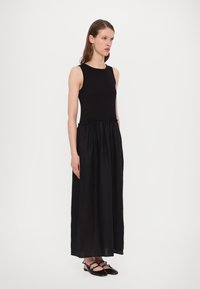 Robe maxi sans manches noire avec un corsage ajusté et une taille froncée. Confectionnée en tissu léger et lisse ; à associer avec des chaussures plates noires.