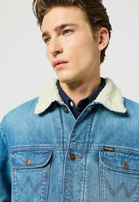 Giacca di jeans azzurro chiaro con colletto in pelliccia sintetica, dotata di chiusure a bottone in rame e due tasche sul petto con cuciture decorative.